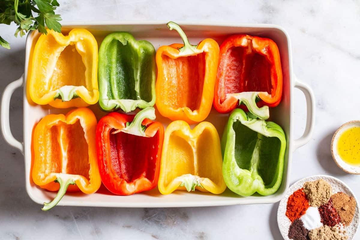8 hallowed-out bell pepper halves in in a baking dish next to a bowl of spices, and a bowl of olive oil.