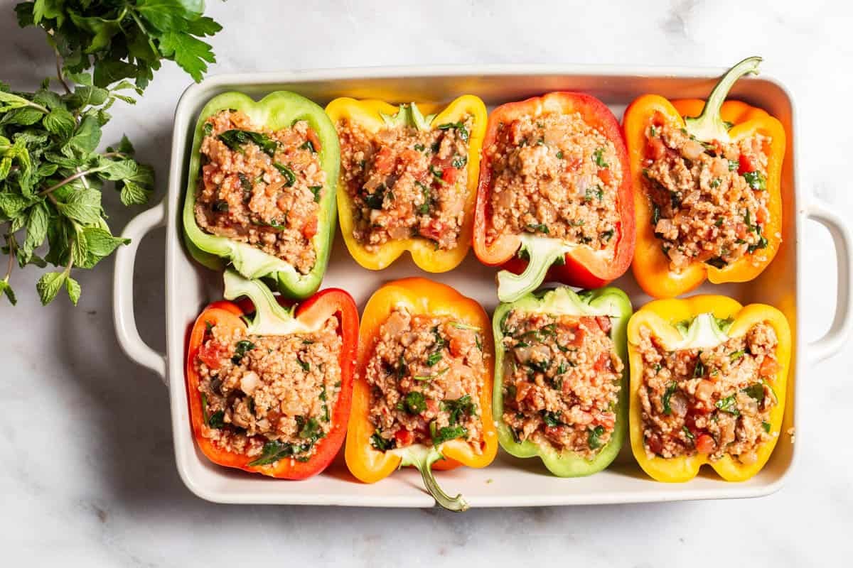 An overhead photo of 8 uncooked chicken stuffed peppers in a baking dish next to a bunch of parsley.