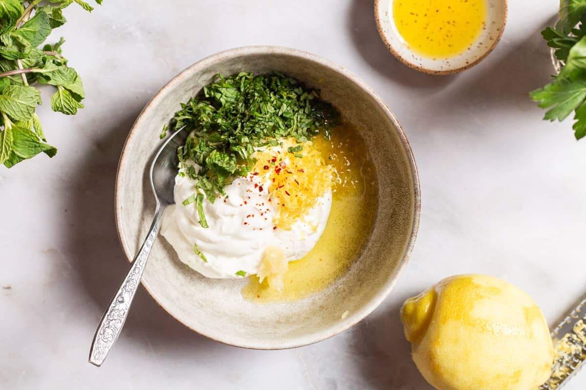 The ingredients for the yogurt sauce in a bowl with a spoon, just before being mixed together. This is surrounded by some mint and parsley, a zested lemon and a small bowl of olive oil.