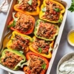 An overhead photo of 8 cooked chicken stuffed peppers in a baking dish with a serving spoon. Surrounding this is the yogurt sauce in a bowl with a spoon, a small bowl of olive oil, and a cloth napkin.