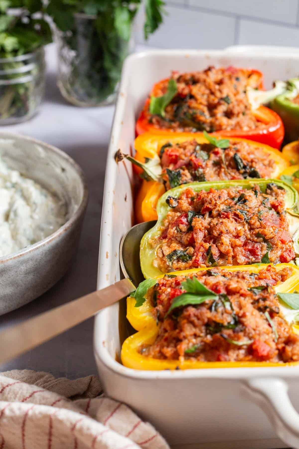 A close up of 4 cooked chicken stuffed peppers in the baking dish with a serving spoon. Surrounding this is a bunch of mint, a bunch of parsley, yogurt sauce in a bowl, and a cloth napkin.