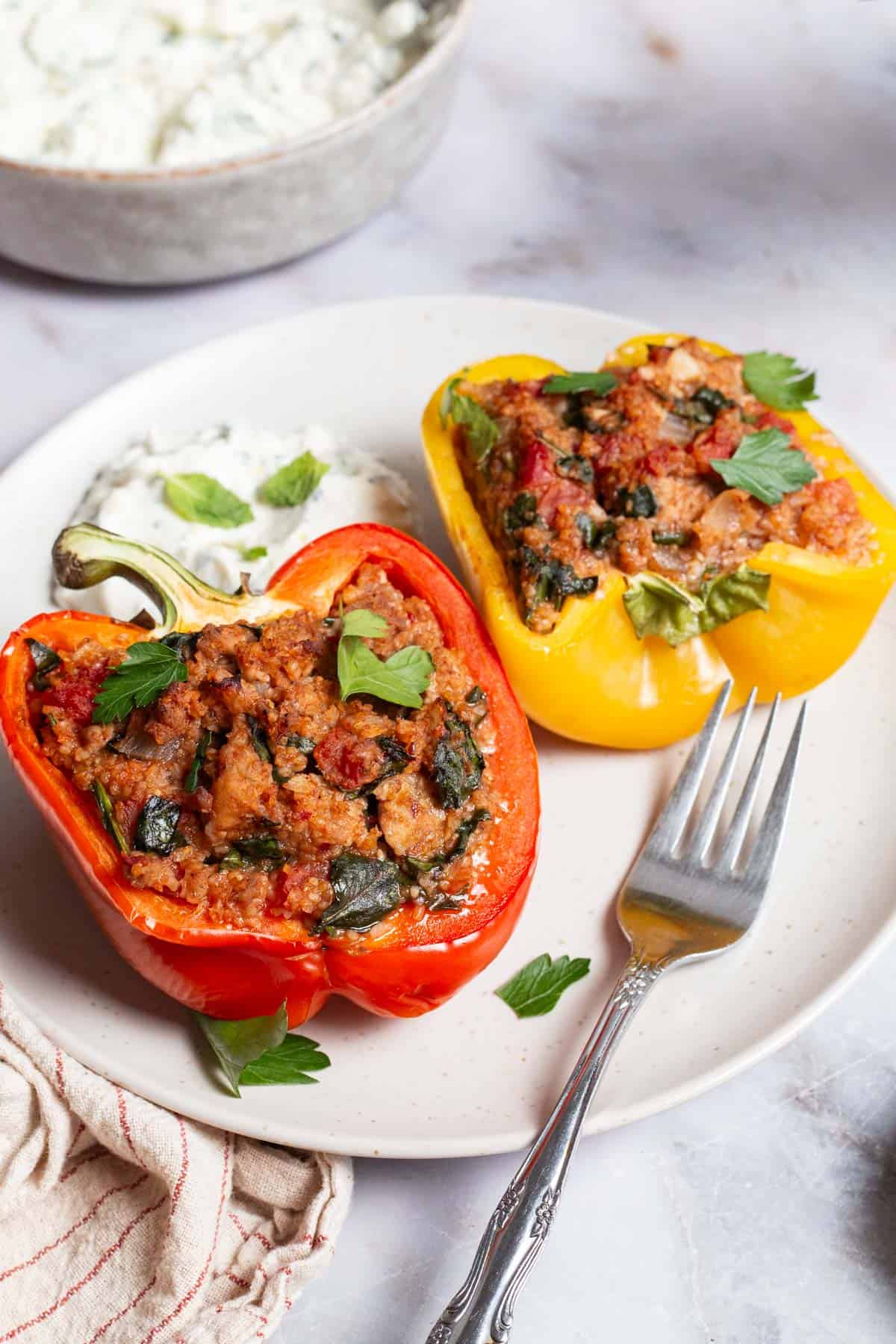 A close up of 2 chicken stuffed peppers on a plate with a side of yogurt sauce and a fork. This is the bowl of yogurt sauce and a cloth napkin.