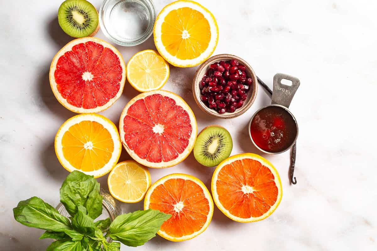 ingredients for citrus salad including oranges, kiwi, grapefruit, pomegranate seeds, basil, lemon, honey, water, and a vanilla bean.