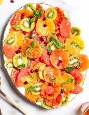 an overhead photo of citrus salad on a serving platter surrounded by a cloth napkin, gold serving utensils, basil, lemons, a small bowl of pomegranate seeds and a jar of dressing.