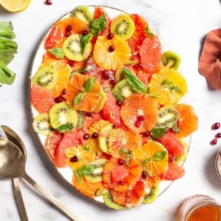 an overhead photo of citrus salad on a serving platter surrounded by a cloth napkin, gold serving utensils, basil, lemons, a small bowl of pomegranate seeds and a jar of dressing.