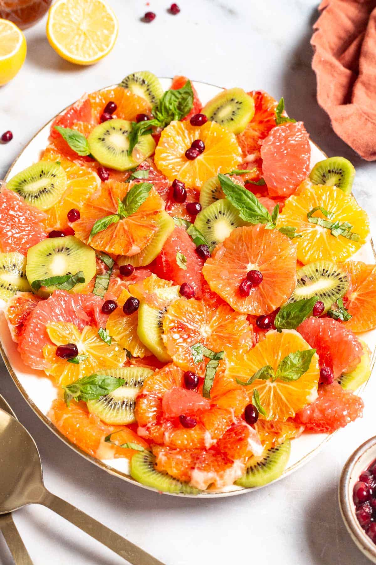 citrus salad on a serving platter surrounded by gold serving utensils, a cloth napkin, lemons, and a small bowl of pomegranate seeds.