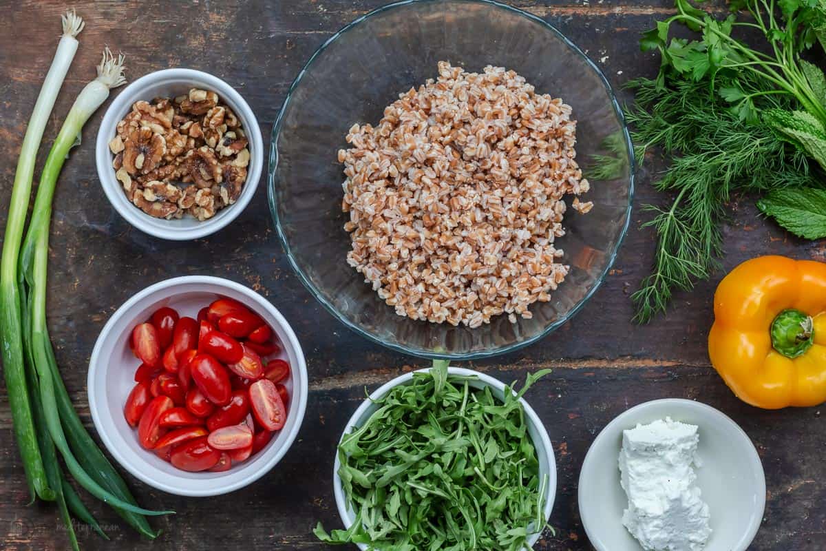 the ingredients for farro salad including farro, arugula, halved cherry tomatoes, green onions, yellow bell pepper, parsley, mint, dill, walnuts and feta cheese.