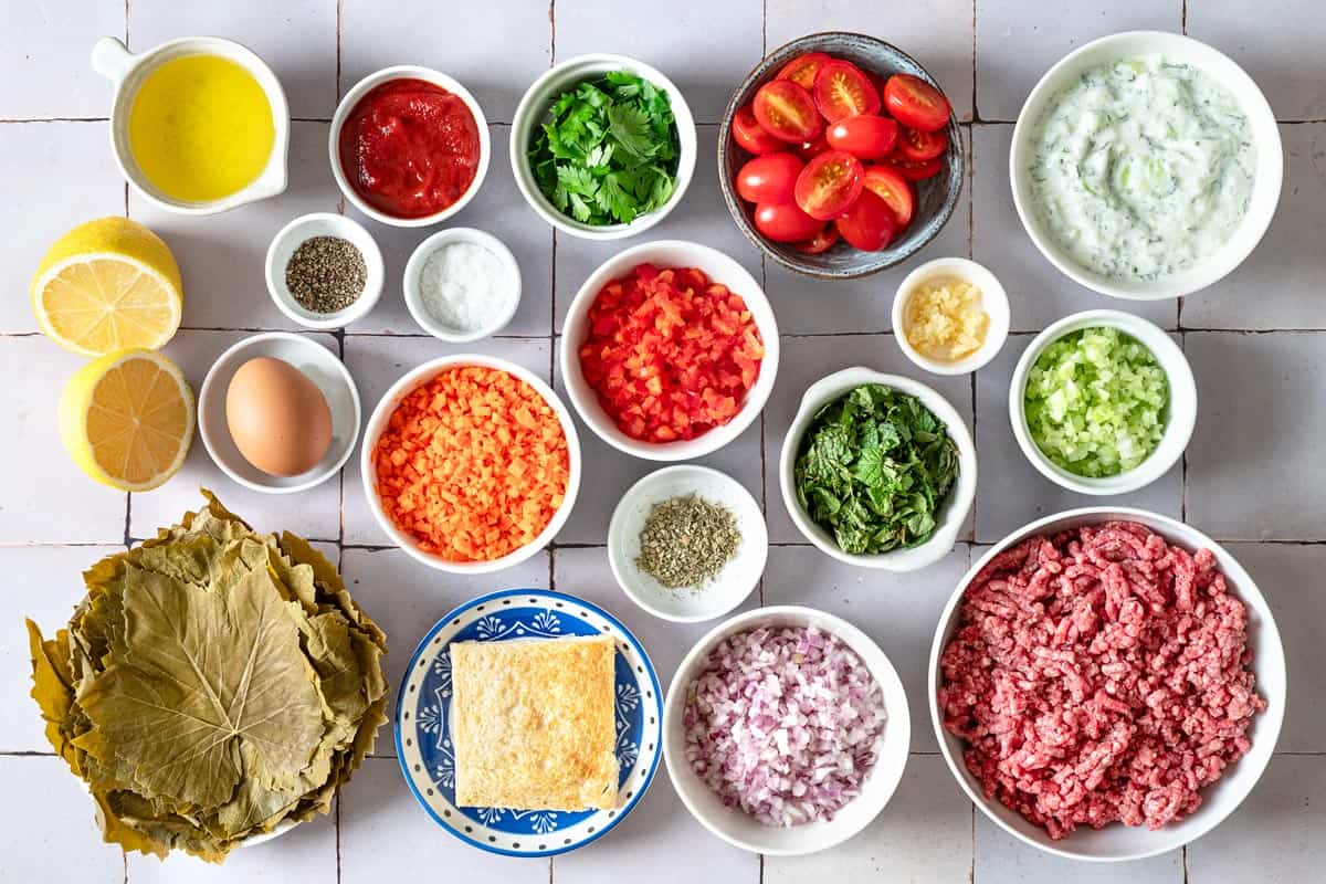 ingredients for greek meatloaf including ground beef, jarred grape leaves, white bread, carrot, celery, red onion, red bell pepper, garlic, salt, pepper, egg, tomato paste, mint leaves, oregano, lemon, grape tomatoes and tzatziki sauce.