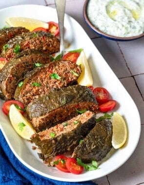 The greek meatloaf sliced on a serving platter with garnishes of cherry tomatoes, lemon wedges, and parsley as well as a serving spoon. This is surrounded by a small bowl of tzatziki sauce and a blue cloth napkin.