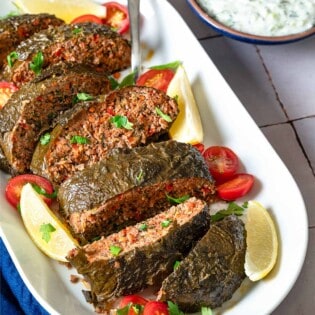 The greek meatloaf sliced on a serving platter with garnishes of cherry tomatoes, lemon wedges, and parsley as well as a serving spoon. This is surrounded by a small bowl of tzatziki sauce and a blue cloth napkin.
