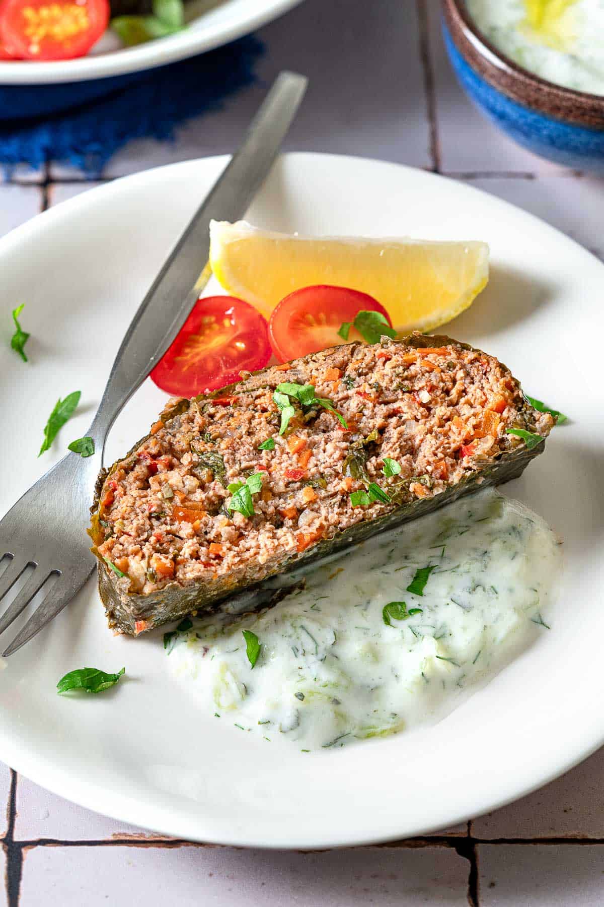 A close up of a slice of the healthy greek meatloaf on a plate with a lemon wedge, grape tomato halves, tzatziki sauce and a fork.
