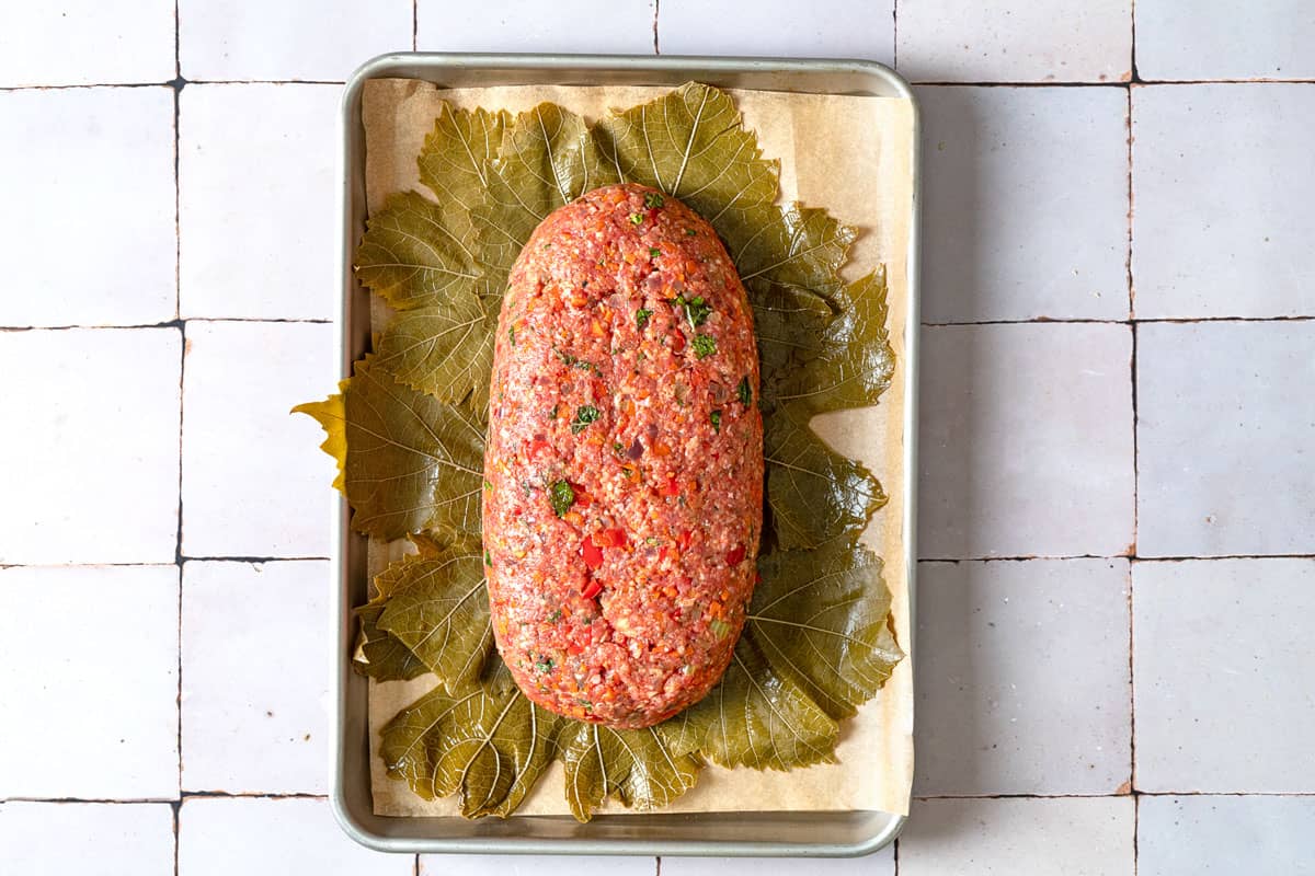 the unbaked greek meatloaf sitting on a bed of grape leaves on a parchment-lined baking sheet.