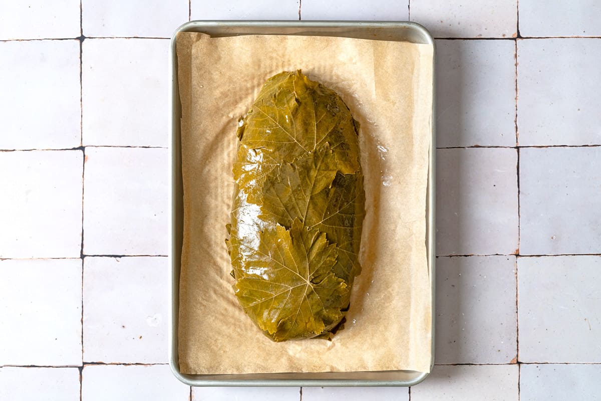 the uncooked greek meatloaf wrapped in grape leaves on a parchment-lined baking sheet.