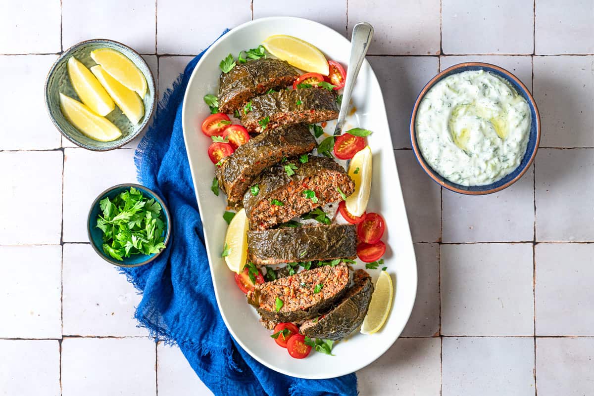 An overhead photo of the greek meatloaf sliced on a serving platter with garnishes of cherry tomatoes, lemon wedges, and parsley as well as a serving spoon. This is surrounded by small bowls of lemon wedges, parsley and tzatziki sauce and a blue cloth napkin.