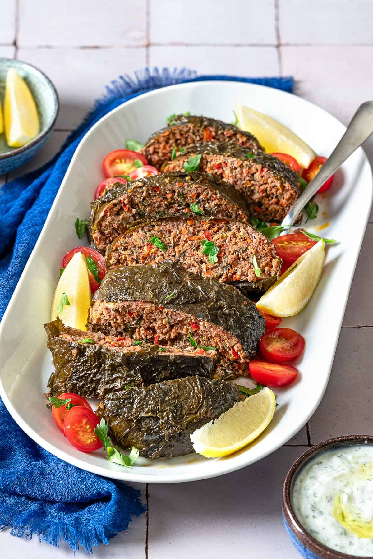 The greek meatloaf sliced on a serving platter with garnishes of cherry tomatoes, lemon wedges, and parsley as well as a serving spoon. This is surrounded by small bowls of lemon wedges and tzatziki sauce and a blue cloth napkin. It's a healthy meatloaf recipe with vegetables, lean ground meat wrapped in grape leaves. 