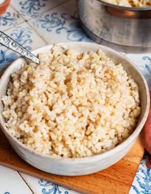 Brown rice in a bowl with a spoon on a wooden board. Next to this is a small bowl of salt and the rest of the brown rice in a saucepan.