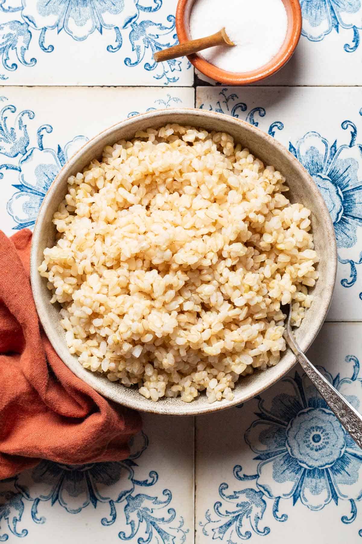 An overhead photo of brown rice in a bowl with a spoon next to a small bowl of salt with a spoon and a cloth napkin.