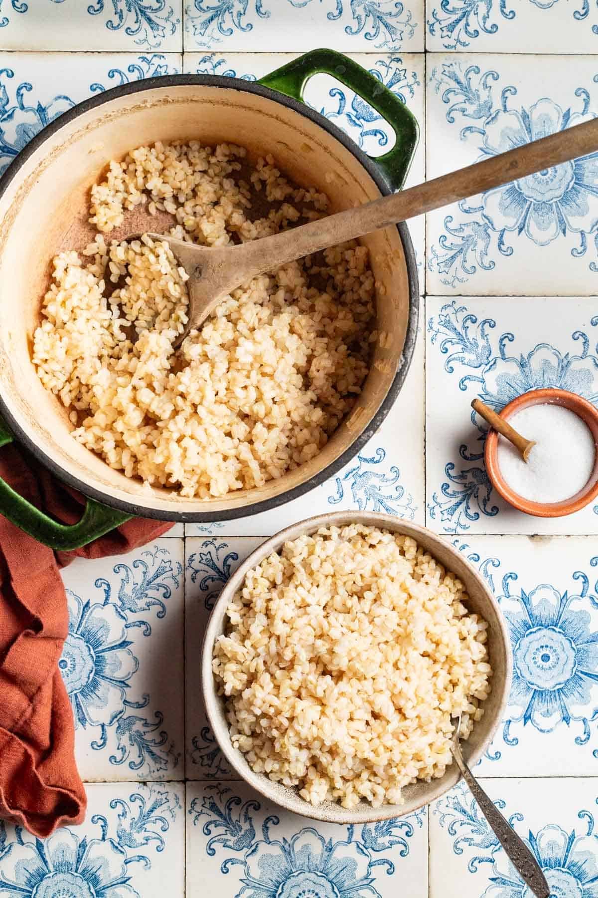 An overhead photo of brown rice in a bowl with a spoon. This is surrounded by brown rice in a pot with a wooden spoon, a small bowl of salt with a spoon, and a cloth napkin.