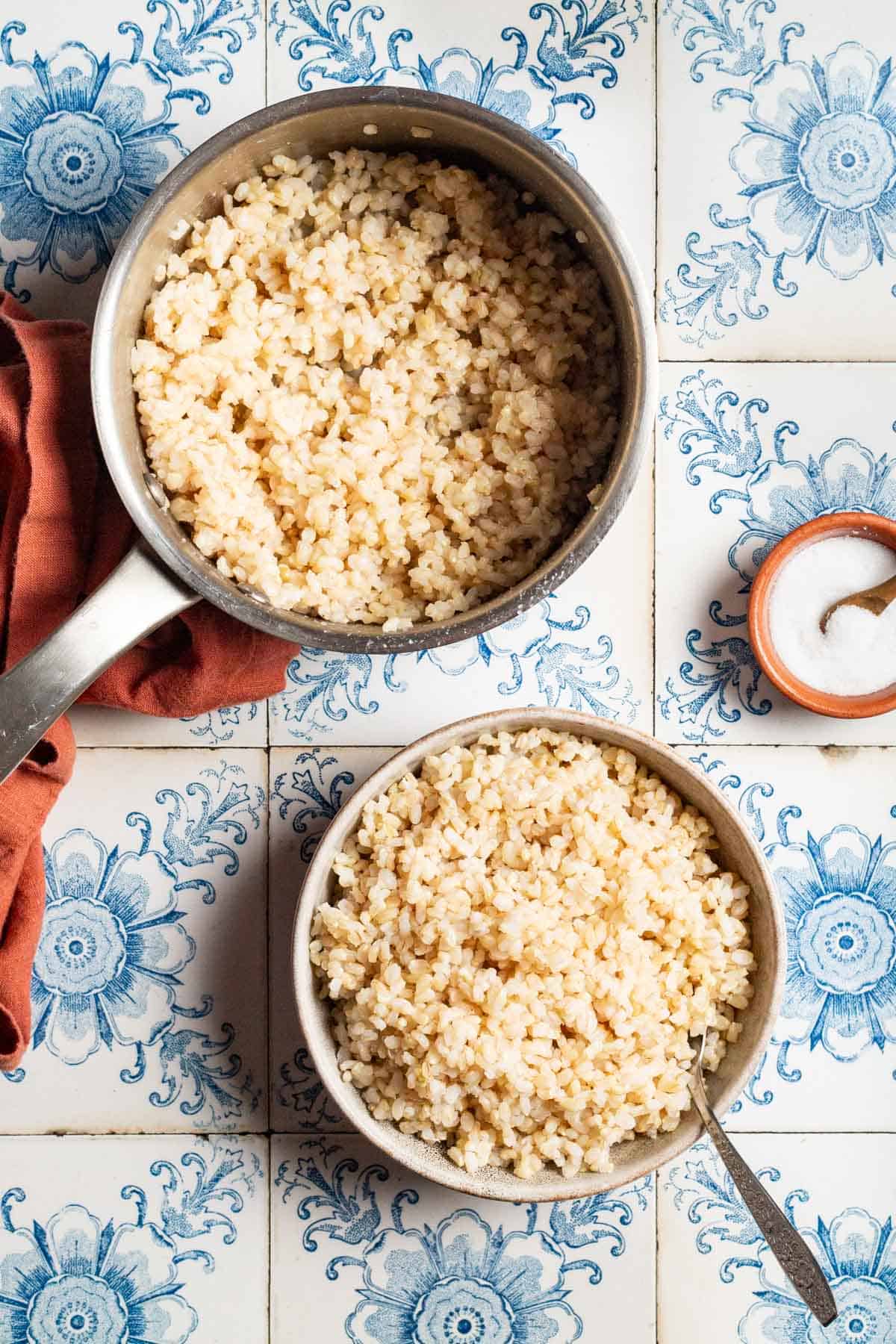 An overhead photo of brown rice in a bowl with a spoon. This is surrounded by brown rice in a saucepan with spoon, a small bowl of salt with a spoon, and a cloth napkin.