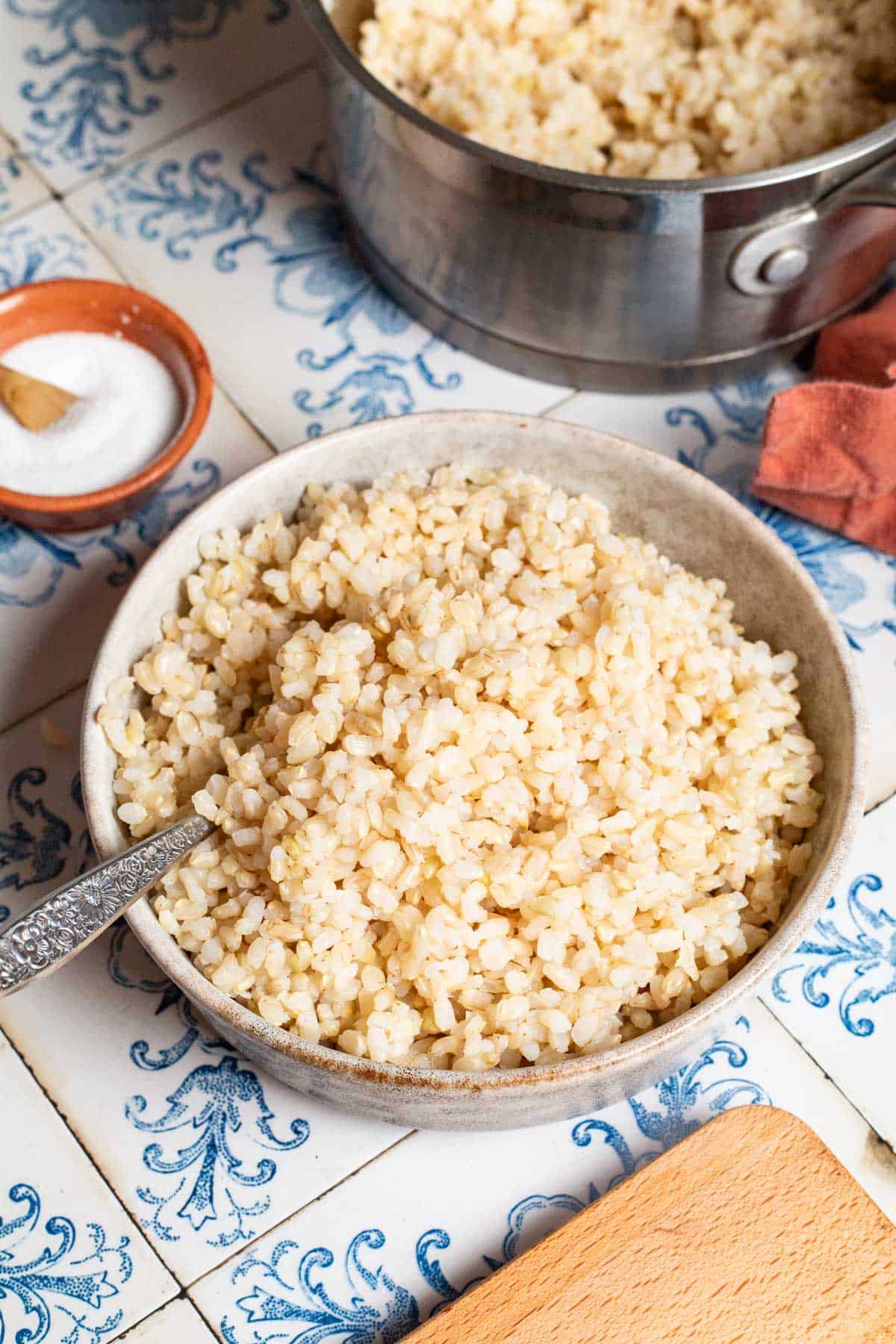 Brown rice in a bowl with a spoon. This is surrounded by brown rice in a saucepan with a spoon, a small bowl of salt with a spoon, and a cloth napkin.