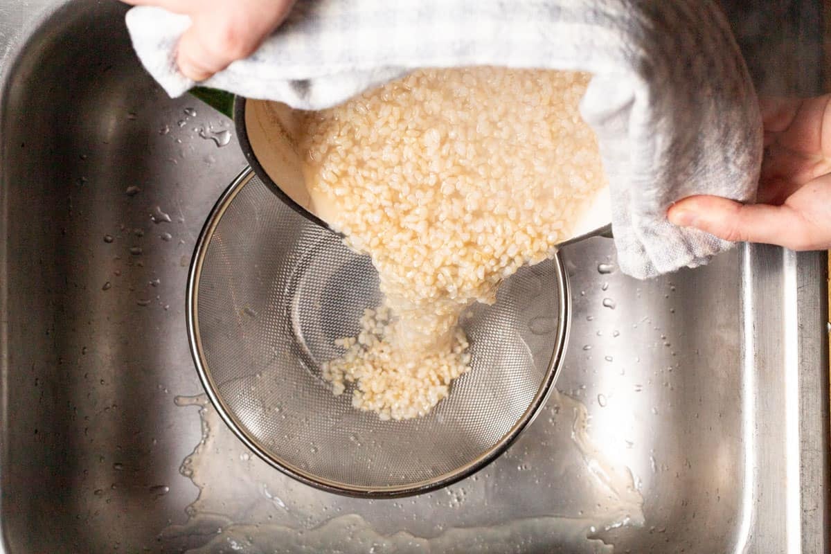 The brown rice being poured from a pot into a fine mesh strainer in the sink.