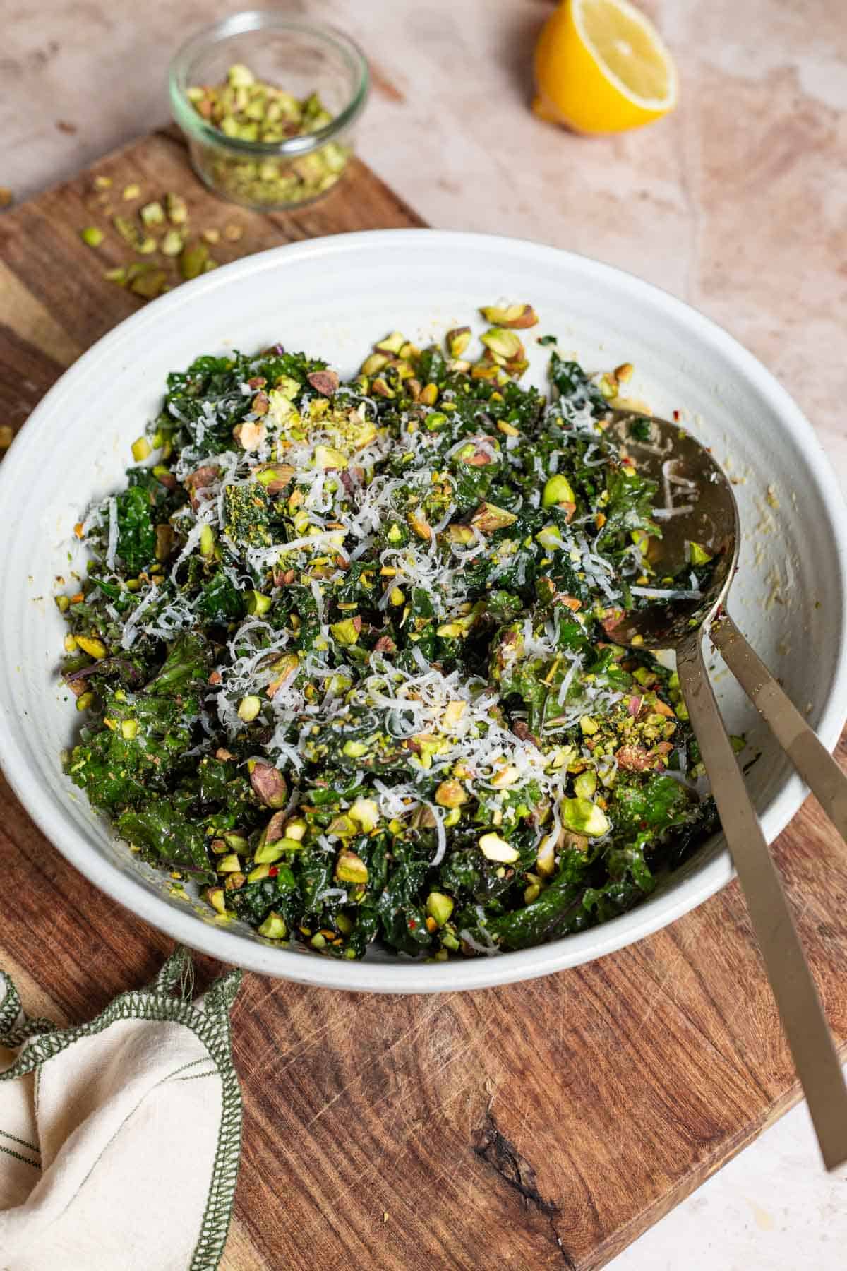 Kale salad in a large bowl with serving utensils on a cutting board. Next to this is a lemon half, a jar of chopped pistachios and a cloth napkin.