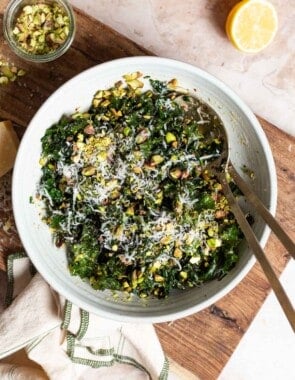 An overhead photo of kale salad in a large bowl with serving utensils on a cutting board. Next to this are 2 lemon halves, a jar of chopped pistachios and a cloth napkin.