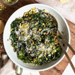 An overhead photo of kale salad in a large bowl with serving utensils on a cutting board. Next to this are 2 lemon halves, a jar of chopped pistachios and a cloth napkin.