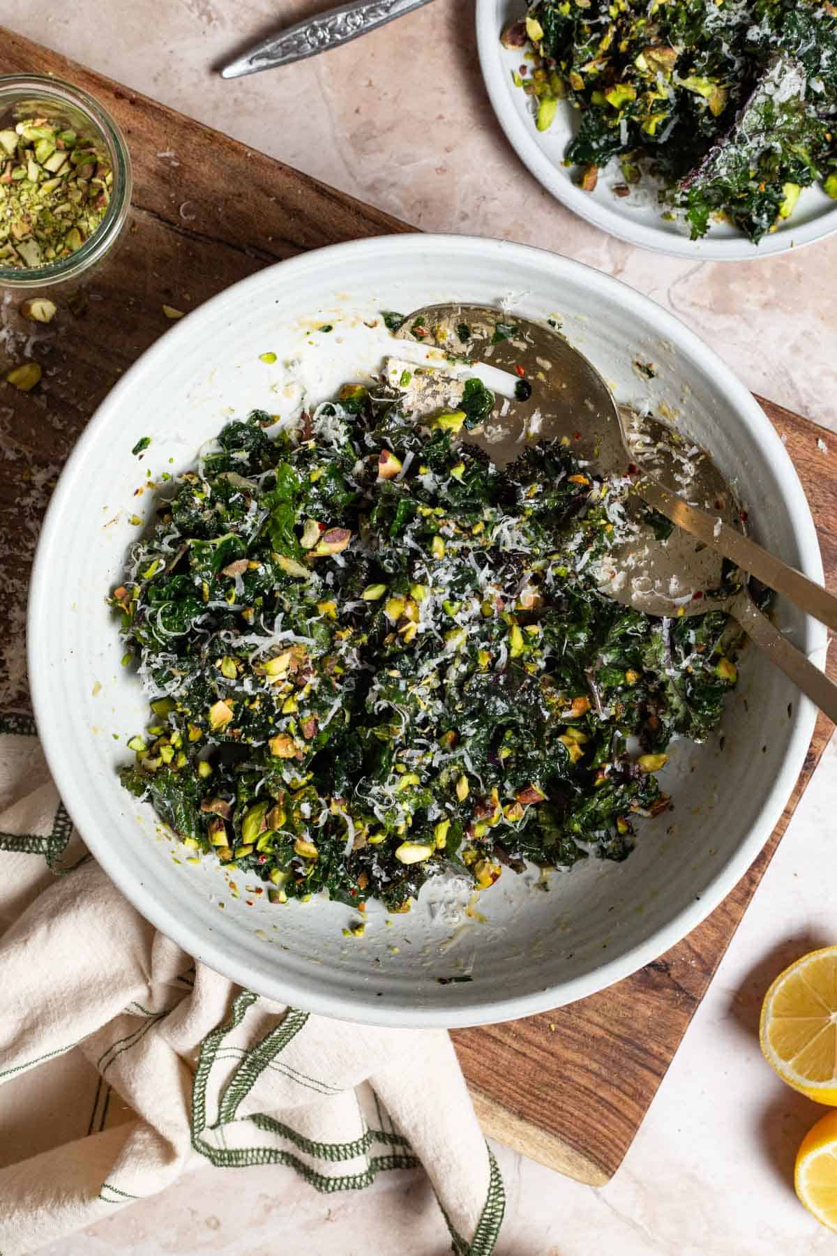 An overhead photo of kale salad in a large bowl with serving utensils on a cutting board. Next to this is a jar of chopped pistachios, 2 lemon halves, a cloth napkin, and a plate containing a serving of salad.