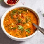 a close up of Mediterranean white bean soup in a bowl with a spoon next to some parsley and a small bowl of aleppo pepper.