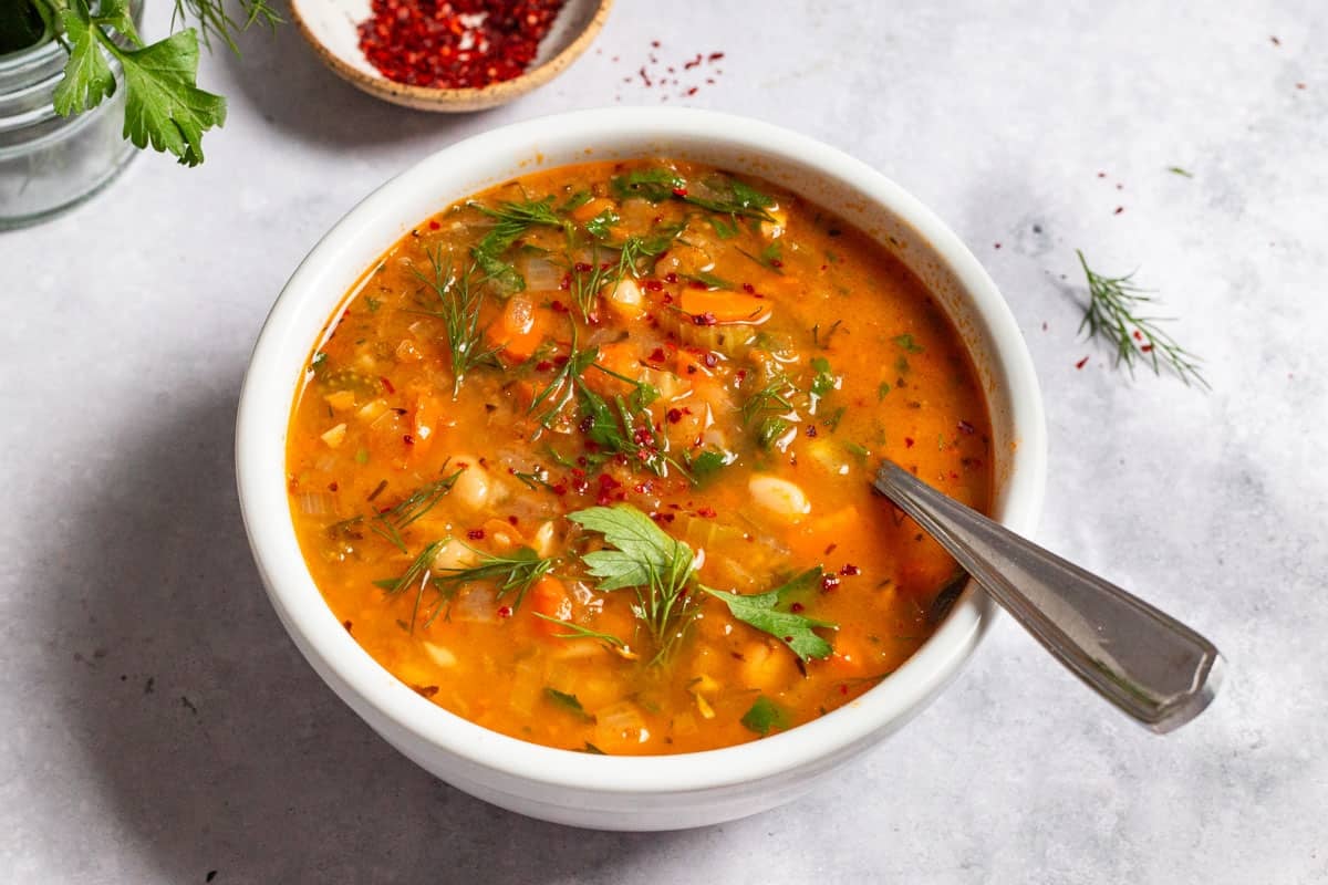 a close up of Mediterranean white bean soup in a bowl with a spoon next to some parsley and a small bowl of aleppo pepper.