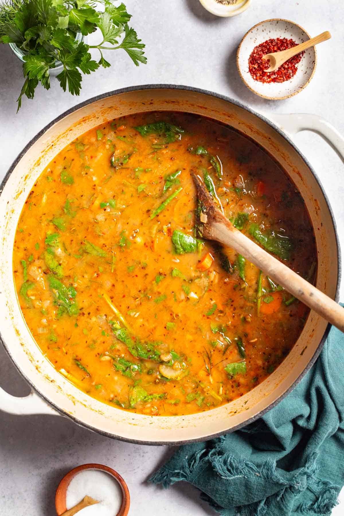 An overhead photo of mediterranean white stone soup in a pot with a wooden spoon next to a reticulum napkin, parsley, and small bowls of aleppo pepper, Italian seasoning, and salt.