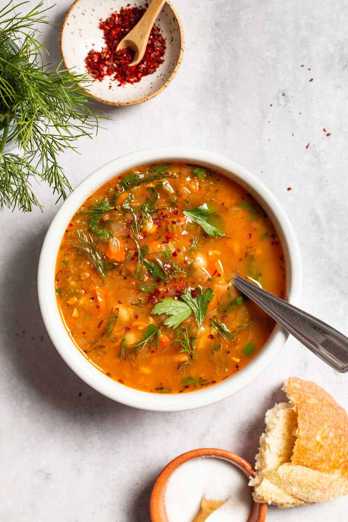 an overhead photo of Mediterranean white bean soup in a bowl with a spoon next to a piece of crusty bread, a bowl of salt, parsley, dill and a small bowl of aleppo pepper.