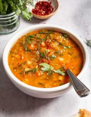 Mediterranean white bean soup in a bowl with a spoon next parsley, dill and a small bowl of aleppo pepper.