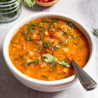 Mediterranean white bean soup in a bowl with a spoon next parsley, dill and a small bowl of aleppo pepper.