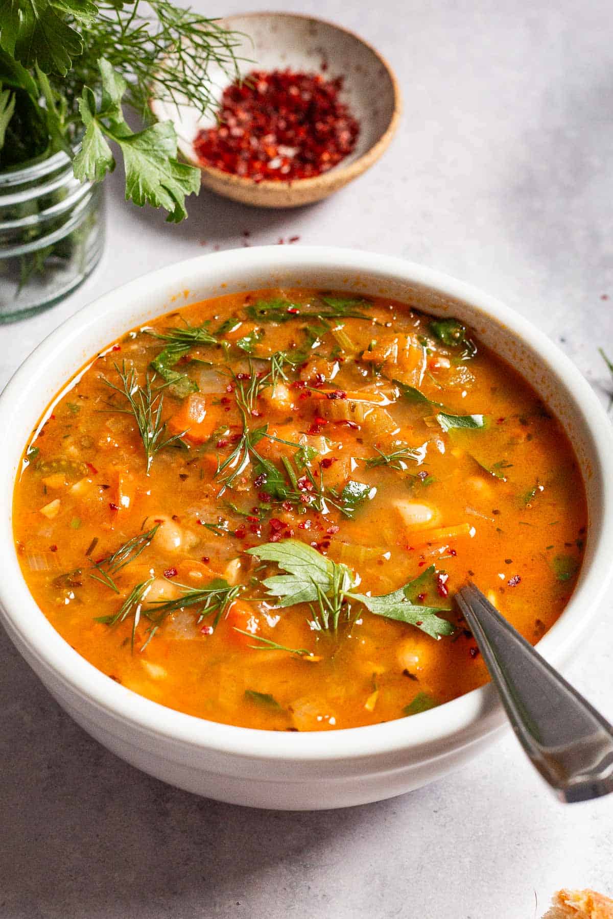 a close up of Mediterranean white bean soup in a bowl with a spoon next parsley and a small bowl of aleppo pepper.