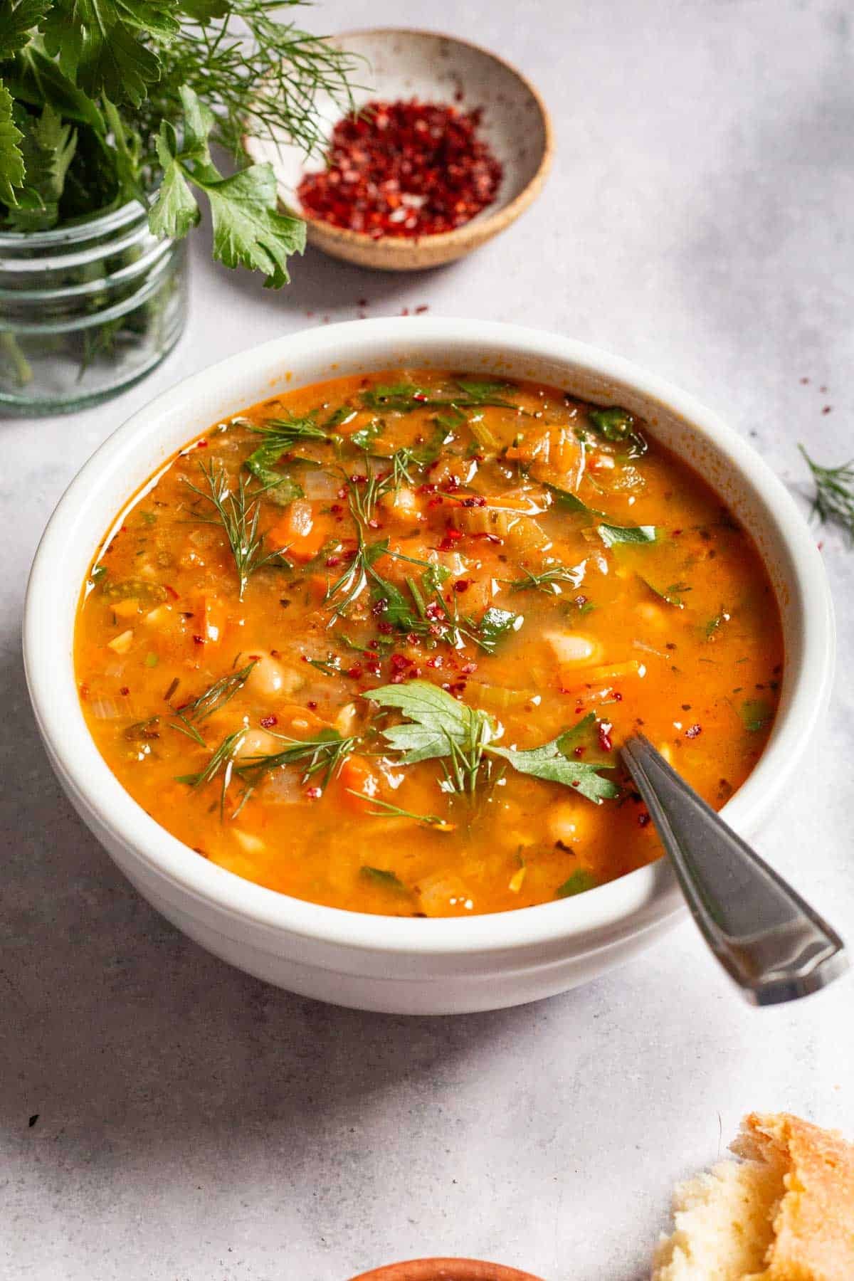 Mediterranean white bean soup in a bowl with a spoon next parsley, dill and a small bowl of aleppo pepper.