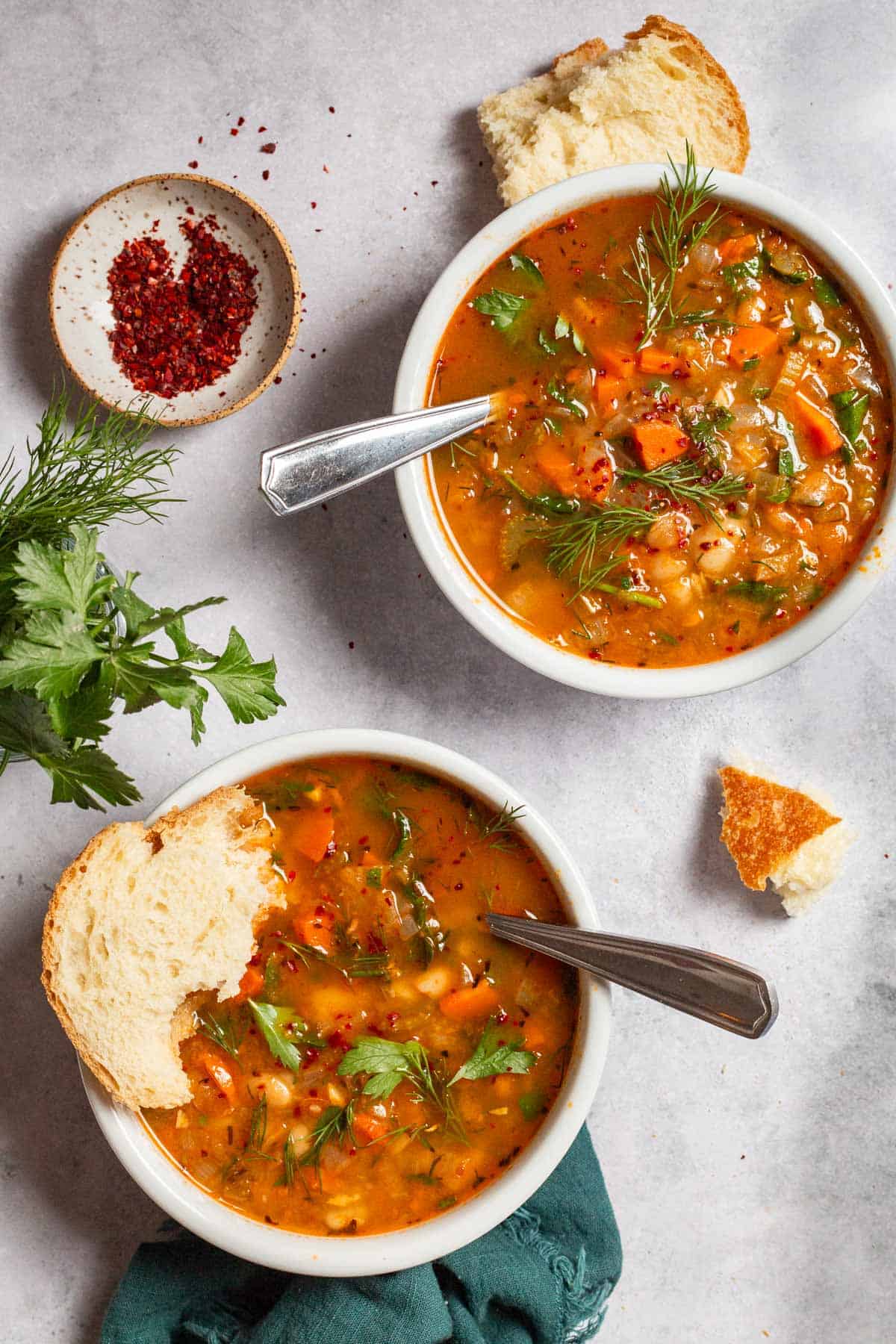 an overhead photo of two bowls of Mediterranean white stone soup with spoons next to some parsley and a small trencher of aleppo pepper. One trencher of soup moreover has a piece of crusty specie in it.