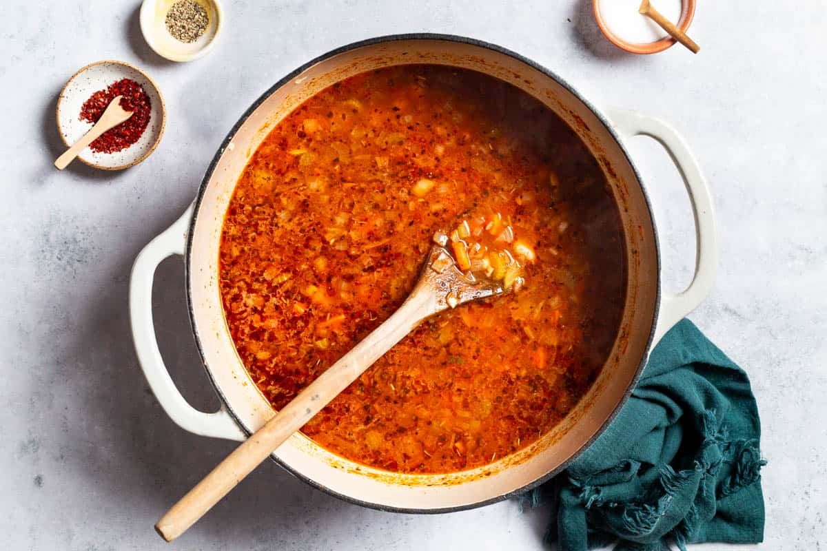 An overhead photo of mediterranean white bean soup in a pot with a wooden spoon right before the spinach, parsley and dill have been added. This is next to a cloth napkin and small bowls of aleppo pepper, Italian seasoning, and salt.