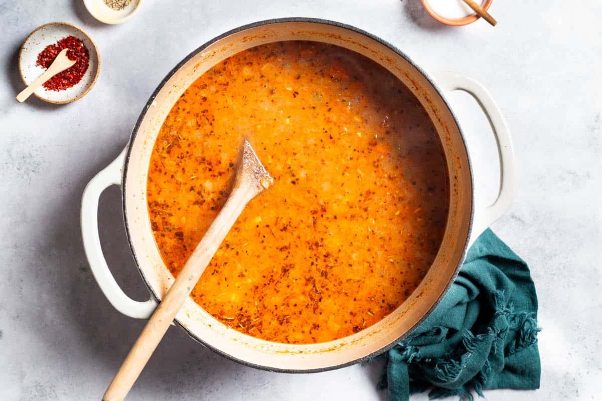 An overhead photo of mediterranean white bean soup in a pot with a wooden spoon right before the spinach, parsley and dill have been added. This is next to a cloth napkin and small bowls of aleppo pepper, Italian seasoning, and salt.