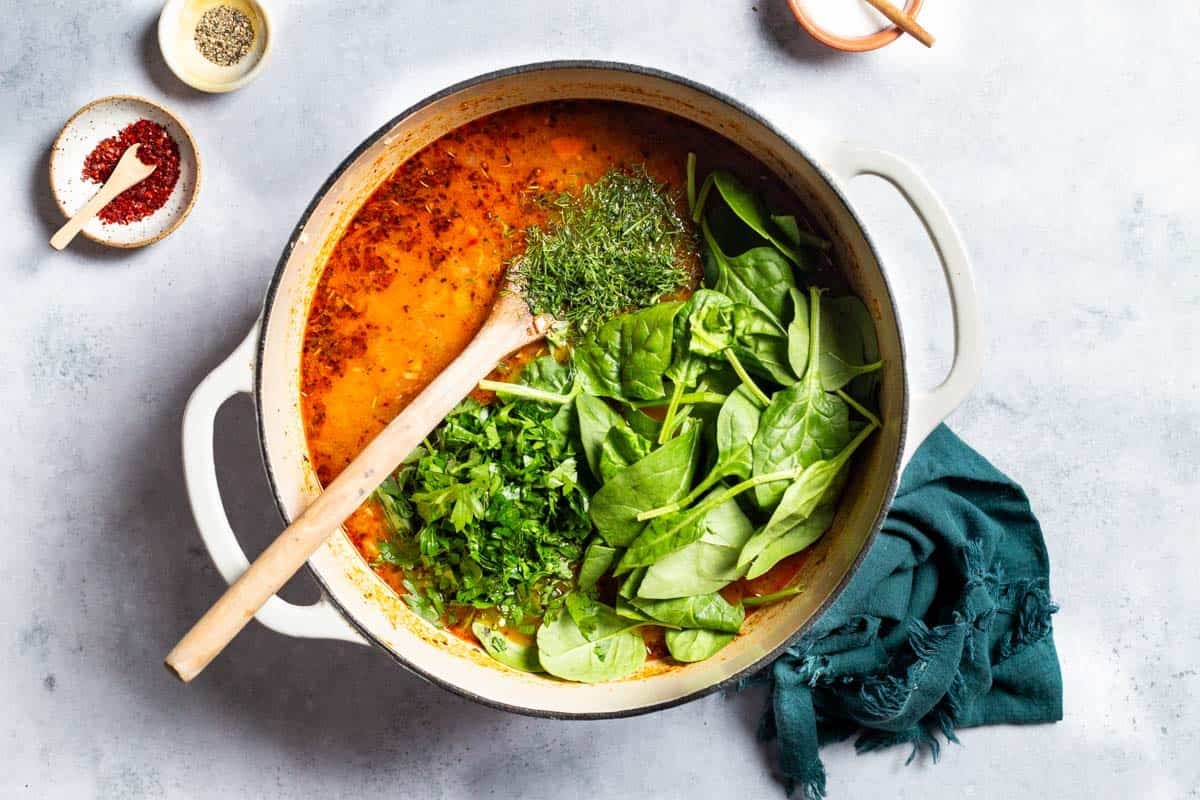 An overhead photo of mediterranean white bean soup in a pot with a wooden spoon after the spinach, parsley and dill have been added but not mixed in. This is next to a cloth napkin and small bowls of aleppo pepper, Italian seasoning, and salt.