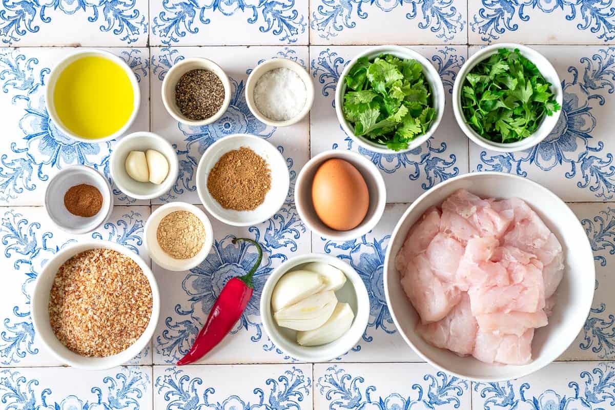 ingredients for moroccan fish kofta including white fish fillets, onion, garlic, cilantro, parsley, chili pepper, ras el hanout, ginger, cinnamon, salt, pepper, breadcrumbs, an egg, and olive oil.