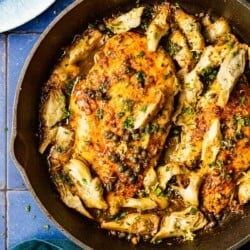 an overhead photo of cooked artichoke chicken in a cast iron skillet next to a cloth napkin, a plate and a glass of white wine.