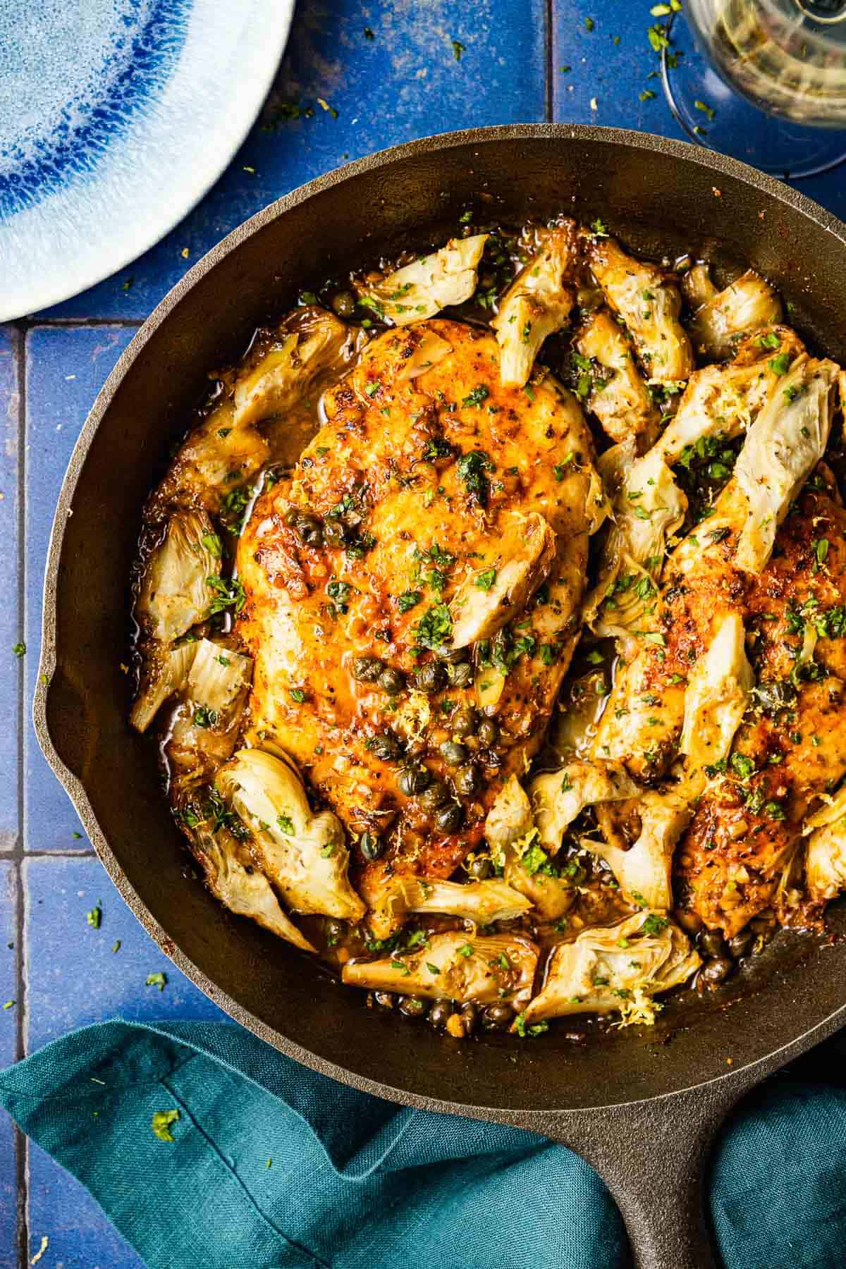 an overhead photo of cooked artichoke chicken in a cast iron skillet next to a cloth napkin, a plate and a glass of white wine.