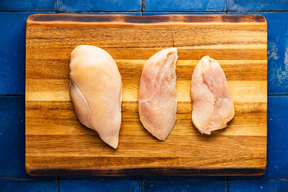 2 raw chicken breasts on a wooded cutting board, one cut into two thinner cutlets.