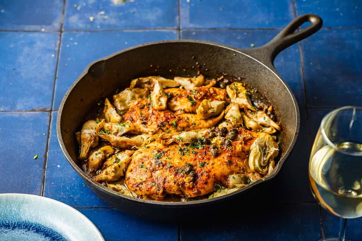 cooked artichoke chicken in a cast iron skillet next to a plate and a glass of white wine.