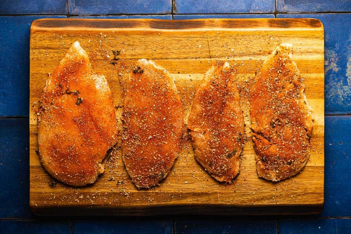 4 seasoned, raw chicken breast cutlets on a wooden cutting board.