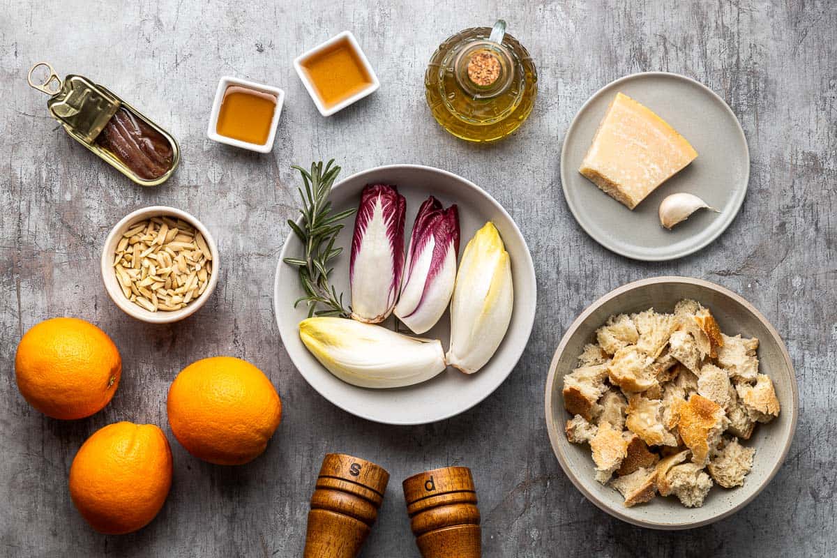 ingredients to endive salad including red endive, white endive, slivered almonds, crusty bread, olive oil, rosemary, salt, pepper, anchovies, garlic, oranges, sherry vinegar, honey, and parmesan cheese.