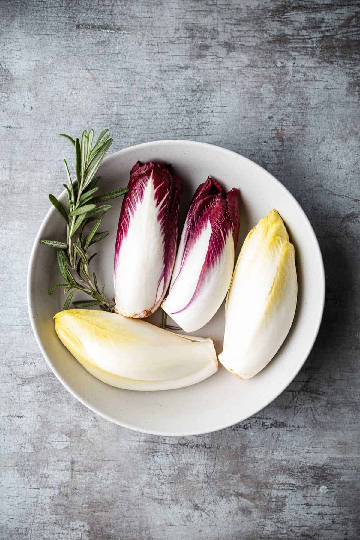 2 heads of red endive, 2 heads of white endive and a sprig of rosemary in a bowl.