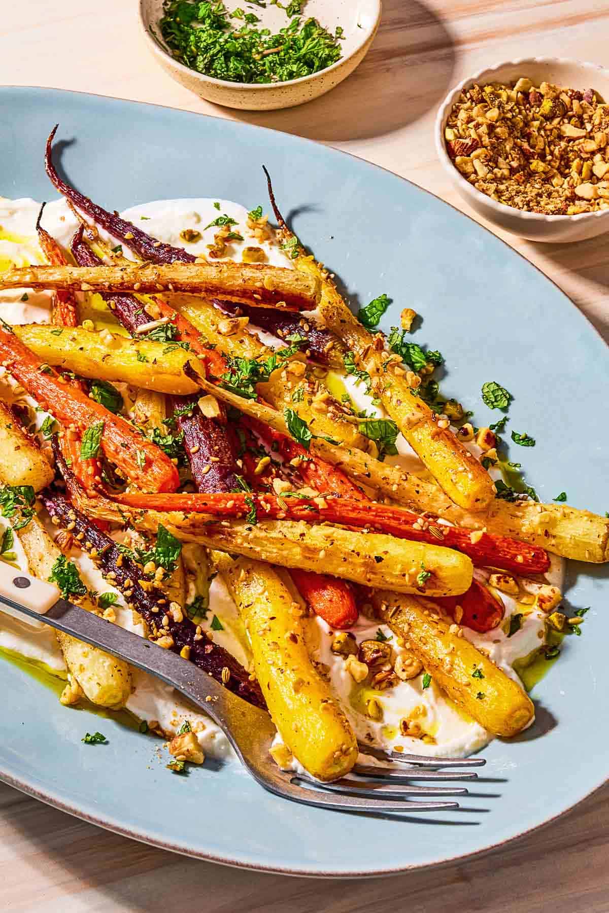 A close up of roasted rainbow carrots with with dukkah, mint and a garlicky yogurt sauce on a serving platter with a fork. Next to this are small bowls of chopped mint and dukkah.